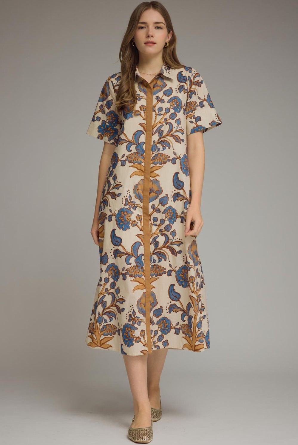 A woman modeling a short-sleeve button-front midi dress with a cream base and an ornate botanical pattern in blue and brown, photographed against a plain light gray studio background.