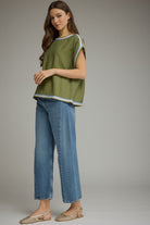 A woman modeling a short-sleeve olive green top with light blue and white striped trim, paired with blue jeans and woven sandals, against a solid gray studio background.