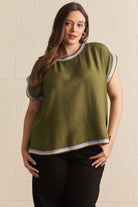 A woman modeling a short-sleeve olive green top with white and gray striped trim on the collar, sleeve cuffs, and hem, paired with black pants against a light beige brick wall.